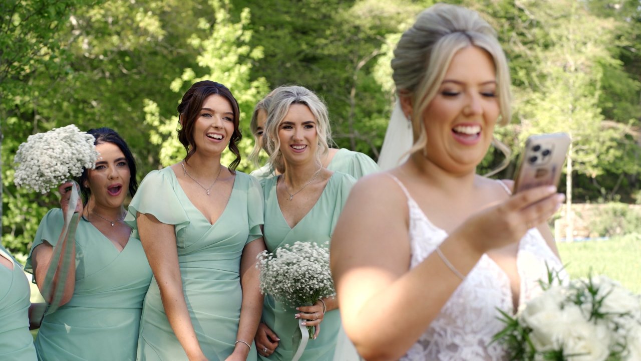 Bride Ciara laughing while looking at a phone with her bridesmaids in sage green dresses.