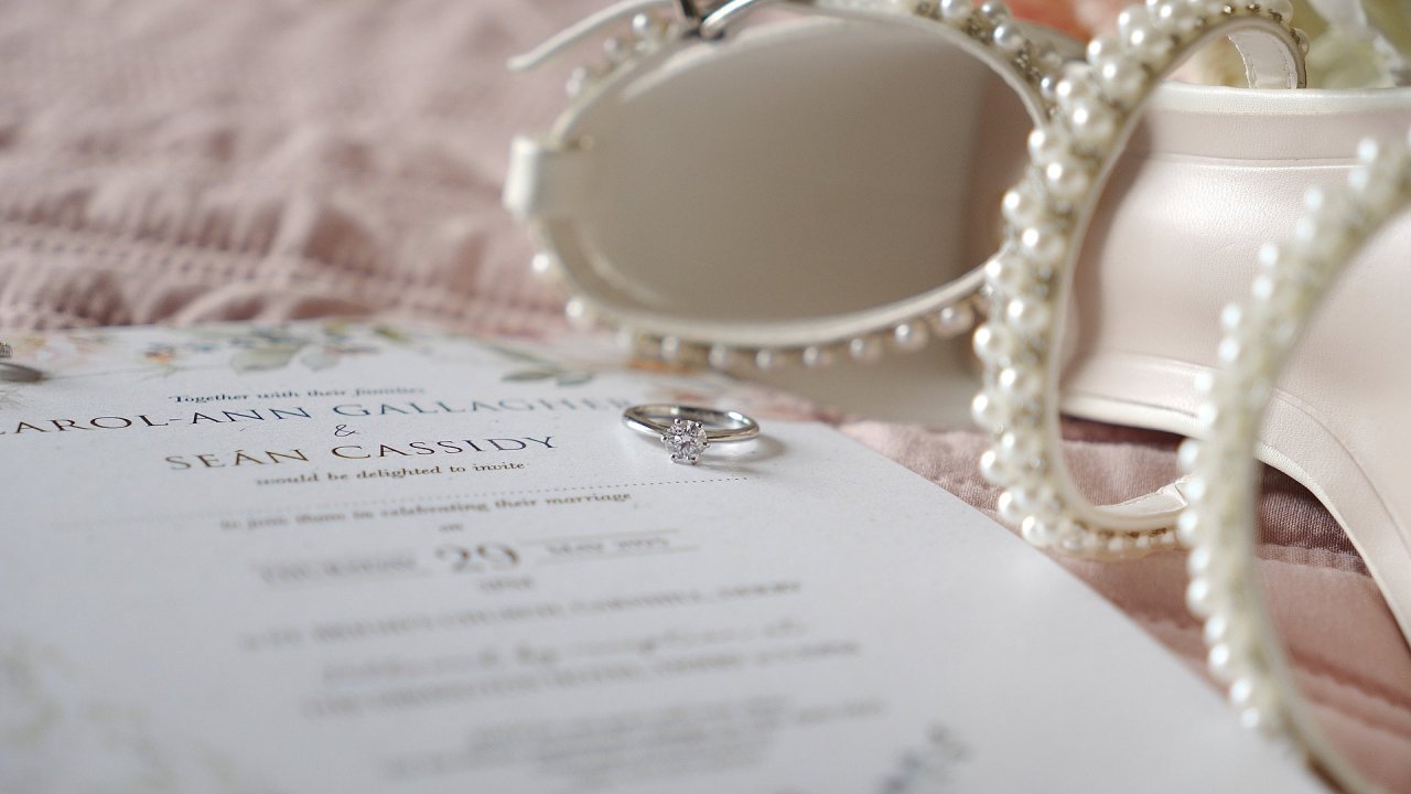 Diamond engagement ring on a wedding invitation for Carol-Ann Gallagher and Sean Cassidy next to pearl-encrusted bridal shoes.
