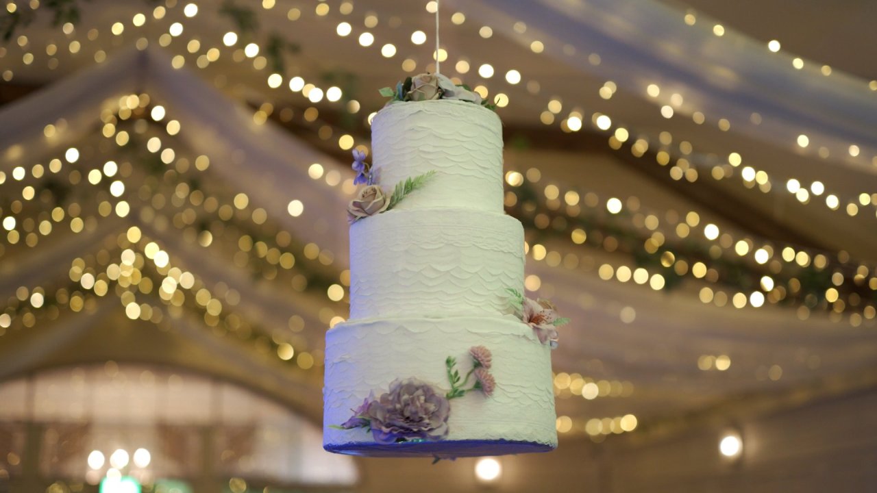 Three-tier white wedding cake piñata suspended from the ceiling with fairy light background.