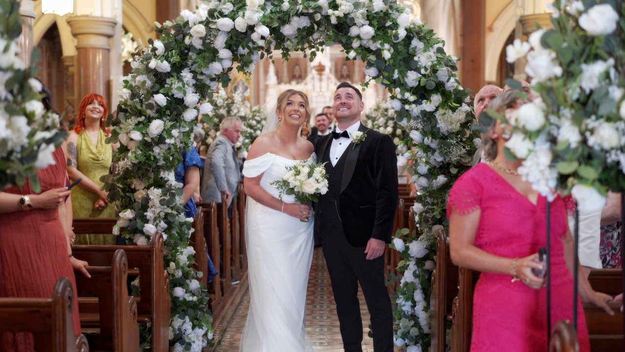 Cora and Conor standing under a lush white floral arch inside the Church of the Immaculate Conception in Strabane.