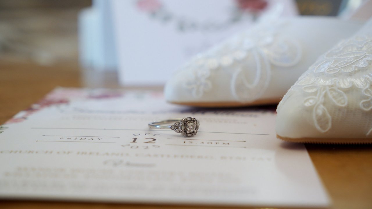 Diamond halo engagement ring placed on a wedding invitation with lace bridal shoes in the background.