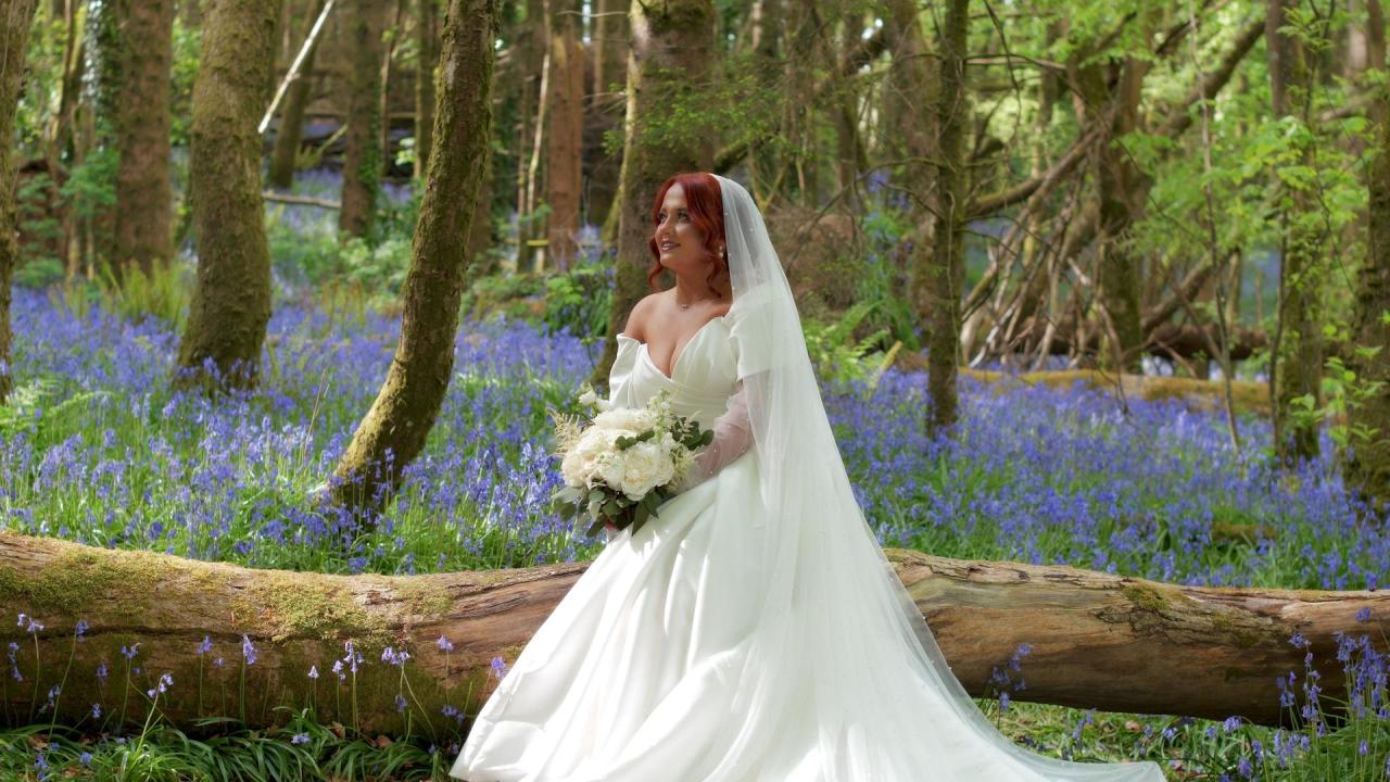 Bride Atlanta in a white off-the-shoulder wedding gown sitting on a log in Rossmore Forest Park bluebell woods.