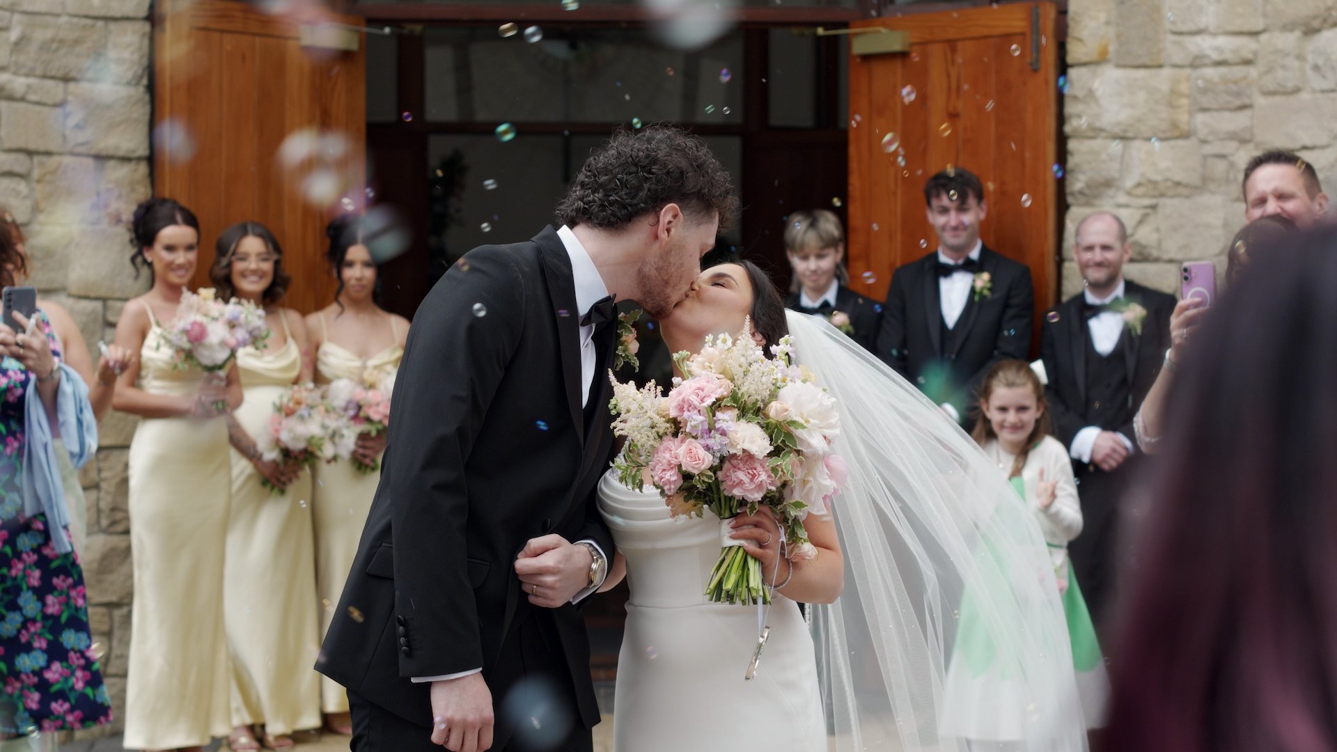 Newlyweds Sophie and Brandon sharing their first kiss outside All Saints Church, Newtowncunningham, surrounded by wedding guests blowing bubbles.