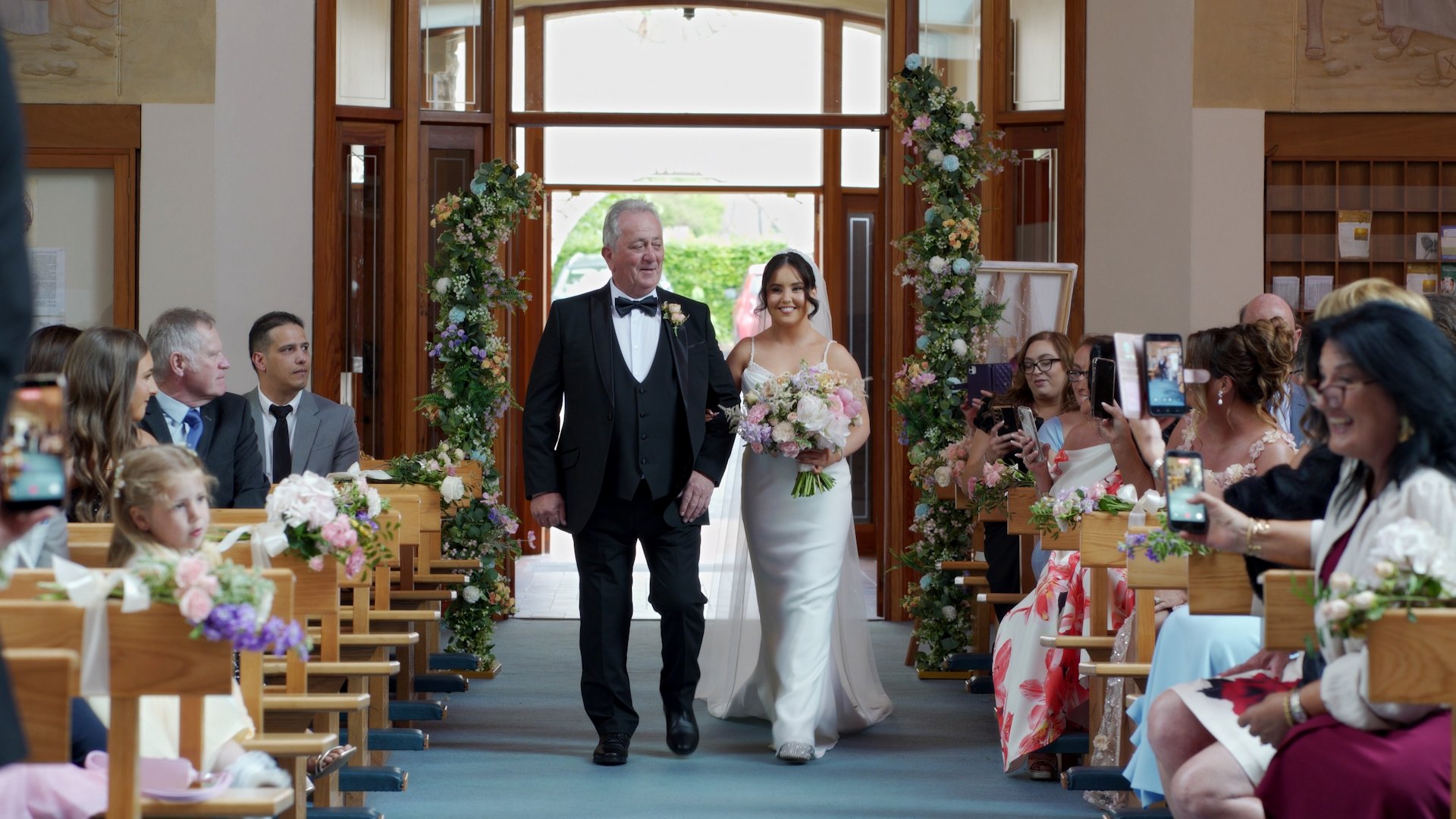 Bride Sophie walking down the aisle at All Saints Church, Newtowncunningham, holding a large pastel pink and white peony bouquet, smiling at guests.