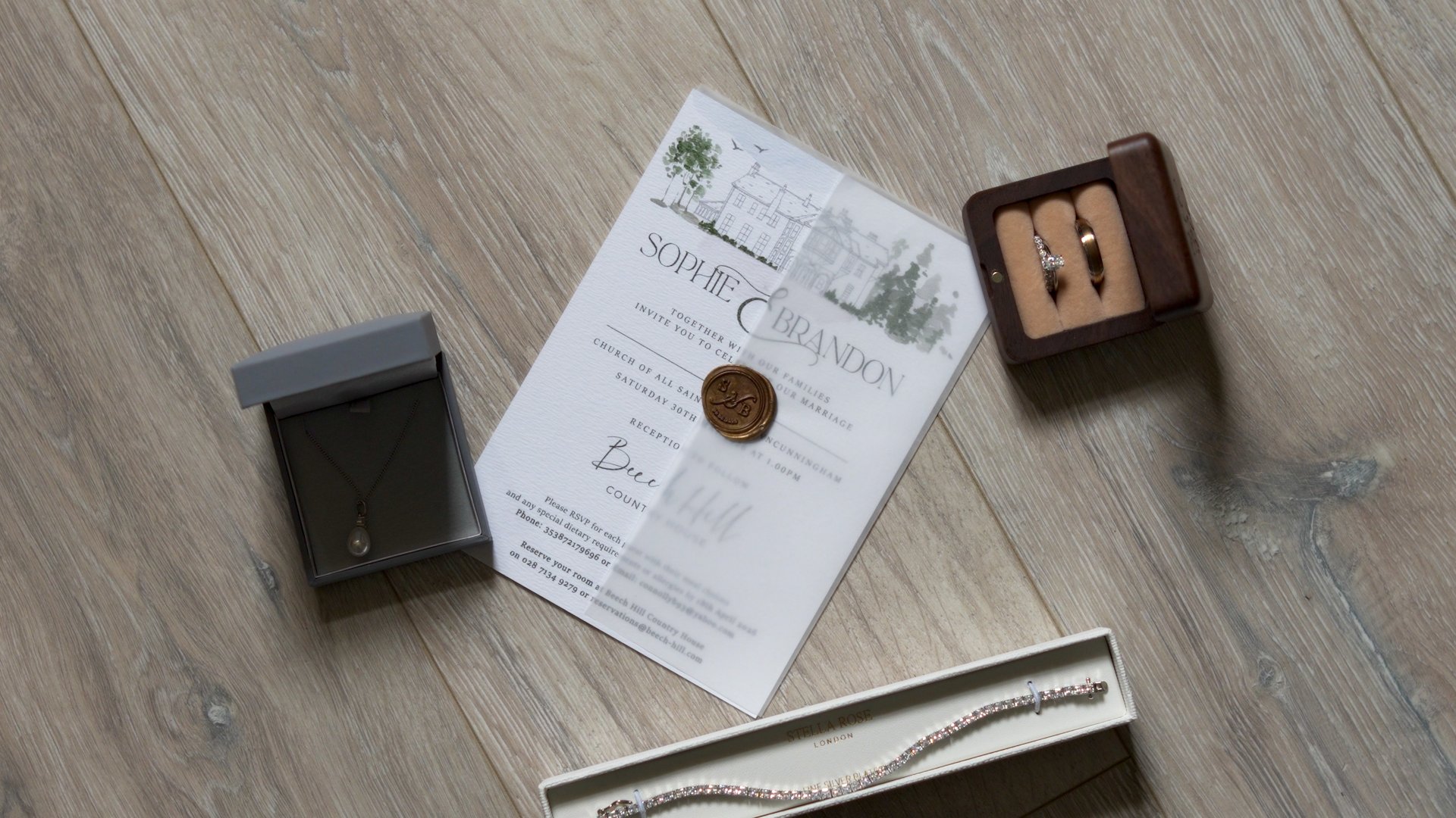 Luxury wedding flat lay on wooden floor featuring Sophie and Brandon's custom vellum invitation with a gold wax seal, a wooden ring box with gold bands, a pearl necklace, and a diamond bracelet.