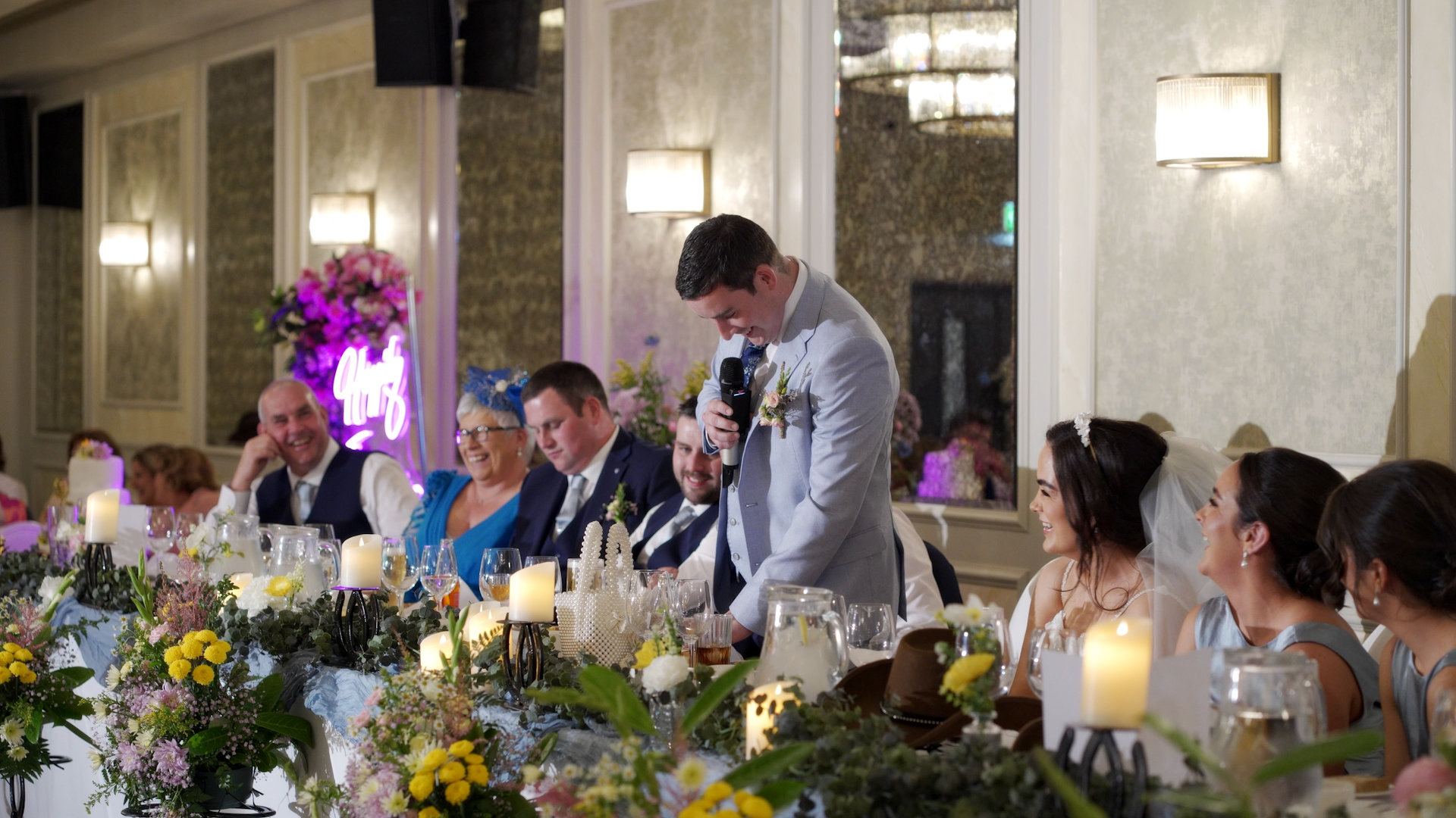 A warm, candid shot of the groom, Jason, speaking at the beautifully decorated top table during the reception speeches, with the bride and bridal party laughing along.