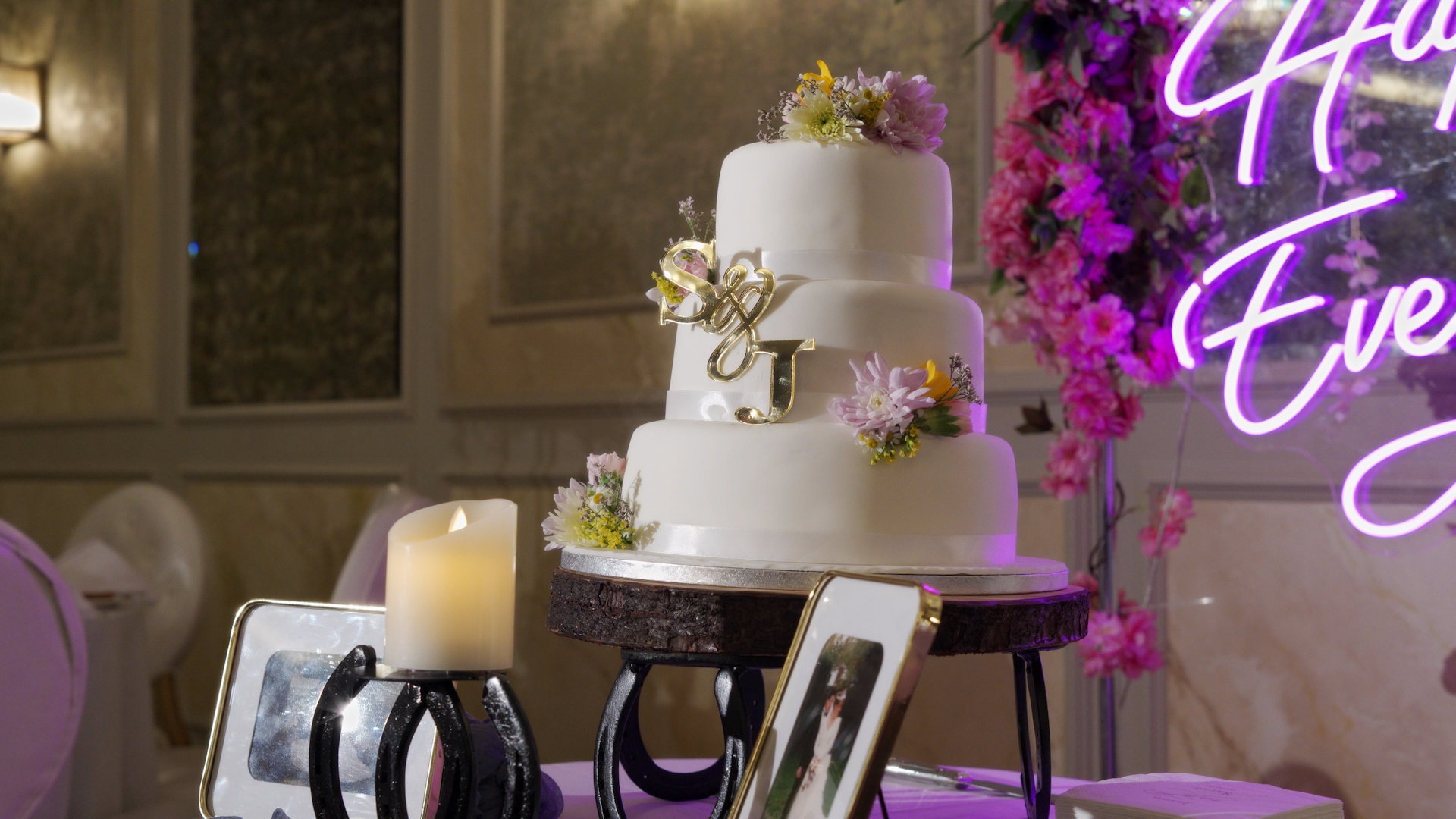 A close-up detail shot of a classic white three-tier wedding cake, adorned with fresh pastel flowers and a custom gold monogram topper featuring the couple's initials, "S & J".