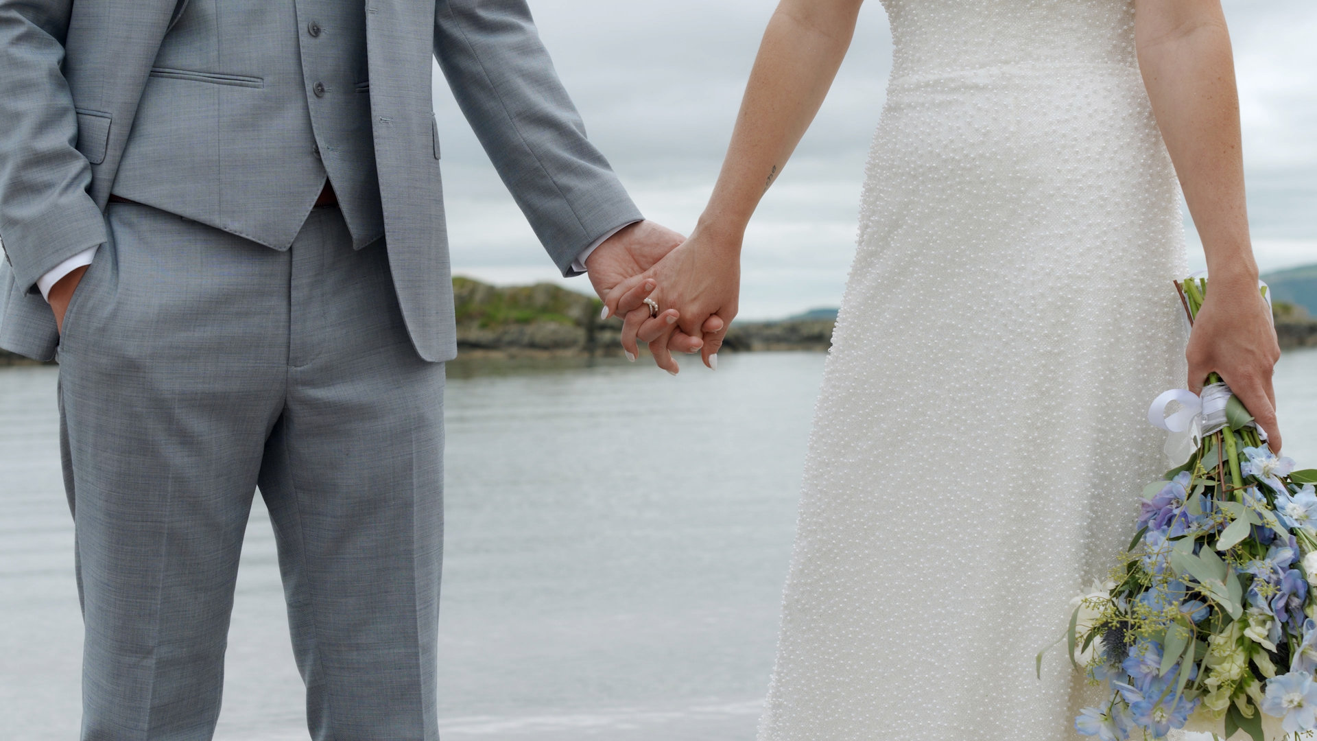 Close-up of bride and groom holding hands, displaying wedding ring and wildflower bouquet with blue delphinium.