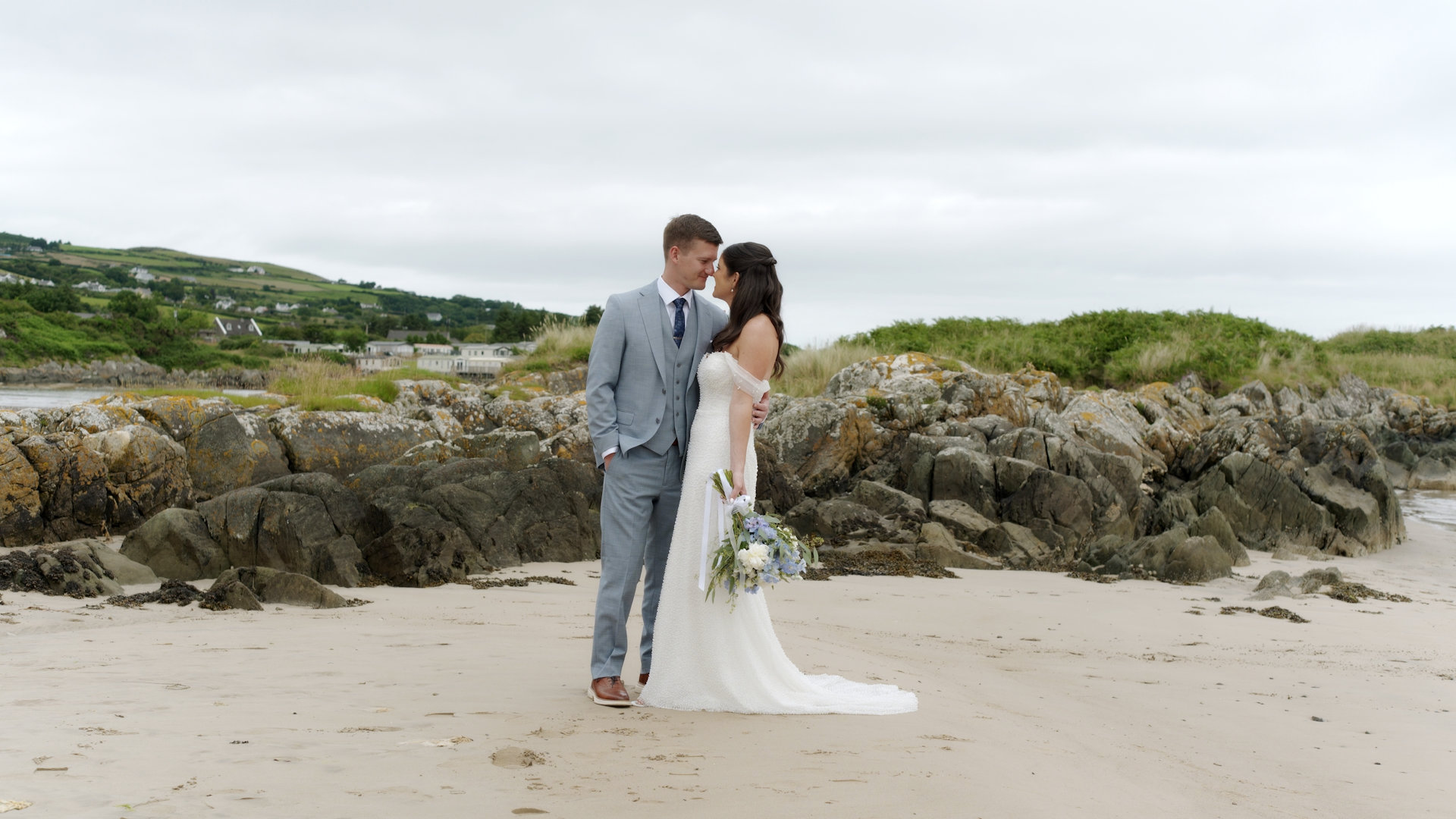 Wide-angle shot of bride Neala Casey and groom Joshua Nichols standing close together on the sandy and rocky shore of Shrove, with the green hills of Donegal in the background.