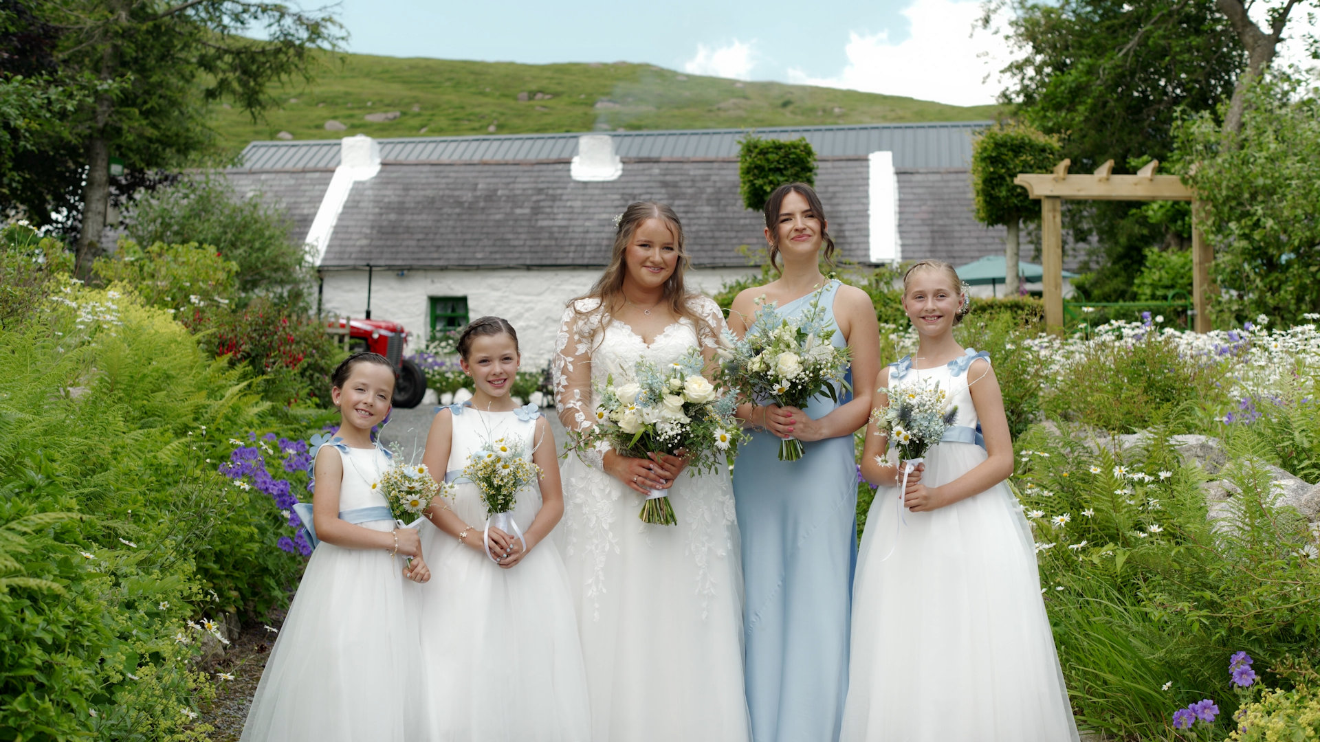 Bride Rebekah with her bridesmaid in a light blue dress and three flower girls holding matching wildflower bouquets outside the cottage.